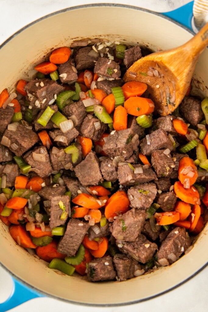 Chunks of browned beef, sliced carrots, celery, and diced onions are being cooked together in a large blue pot with a wooden spoon.