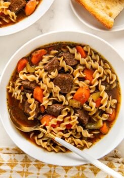 A bowl of beef noodle soup with egg noodles, chunks of beef, carrots, and celery in a rich broth, with a spoon resting inside.