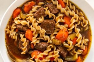 A bowl of beef noodle soup with egg noodles, chunks of beef, carrots, and celery in a rich broth, with a spoon resting inside.