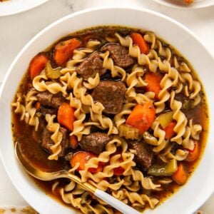 A bowl of beef noodle soup with egg noodles, chunks of beef, carrots, and celery in a rich broth, with a spoon resting inside.