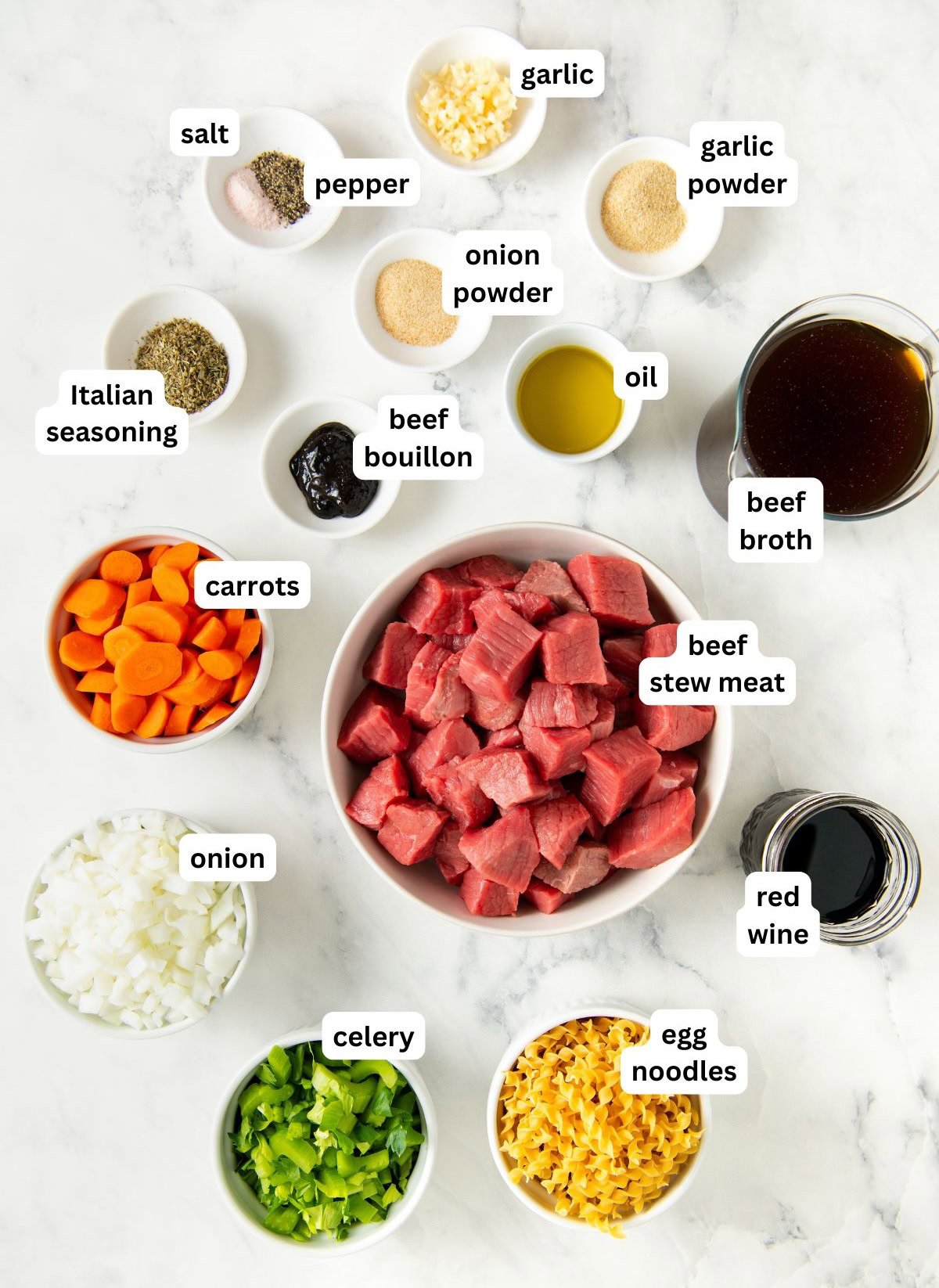 Top-down view of labeled ingredients for beef noodle soup recipe on a white surface, including beef stew meat, onion, celery, carrots, egg noodles, beef broth, beef bouillon, oil, red wine, Italian seasoning, garlic, salt, pepper, onion powder, and garlic powder.