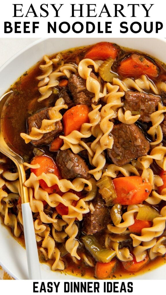 A bowl of beef noodle soup with spiral pasta, chunks of beef, carrots, and celery in a rich broth. A gold and white spoon rests in the soup.