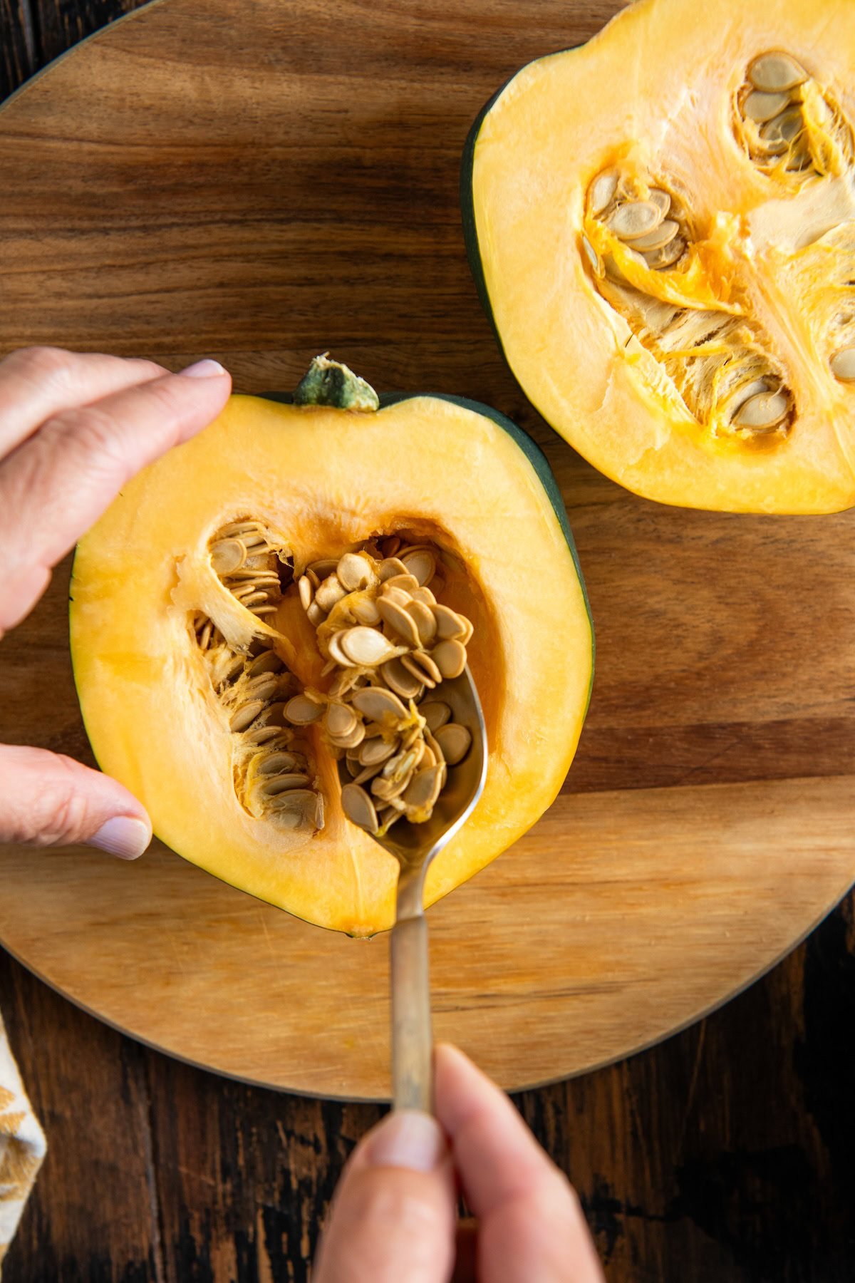 A hand holds a spoon to scoop seeds from a halved acorn squash on a round wooden cutting board.