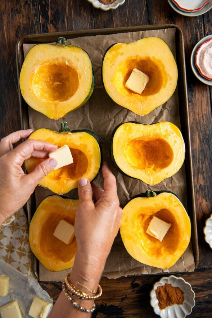 Six halved acorn squashes are on a baking sheet lined with parchment. A person places butter cubes into the squash cavities. Dishes with spices and more butter sit nearby on a rustic wooden table.
