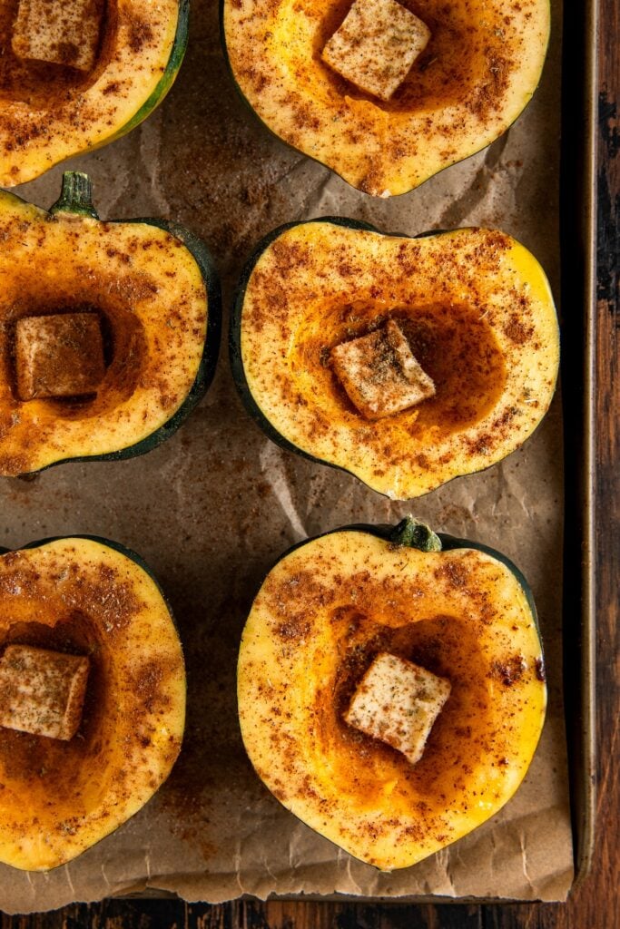 Halved acorn squash on a baking sheet, coated with oil, each filled with a pat of butter and sprinkled with cinnamon and more spices, ready to be roasted.