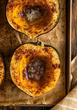 Close-up of two roasted acorn squash halves on a baking sheet, sprinkled with spices and filled with melting butter in the centers.