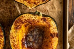 Close-up of two roasted acorn squash halves on a baking sheet, sprinkled with spices and filled with melting butter in the centers.