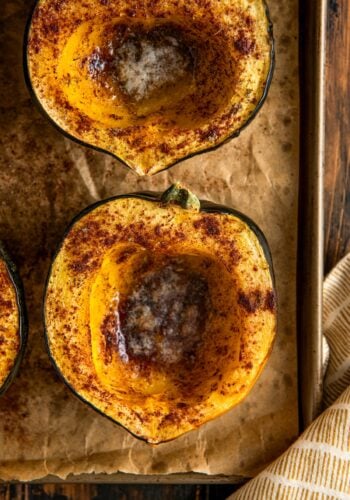 Close-up of two roasted acorn squash halves on a baking sheet, sprinkled with spices and filled with melting butter in the centers.