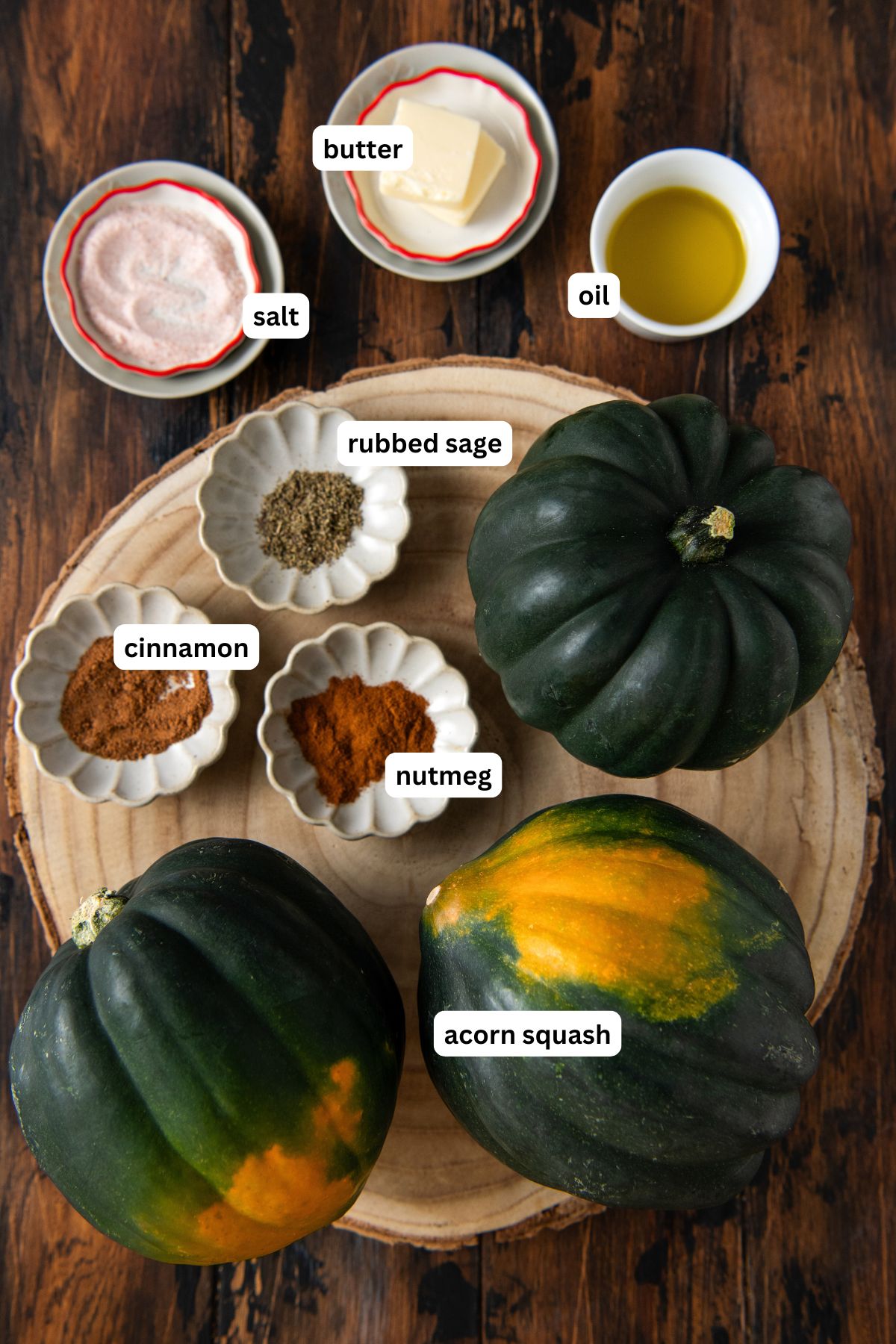 Three acorn squash on a wooden slab, surrounded by small bowls containing butter, oil, salt, rubbed sage, cinnamon, and nutmeg, all labeled.