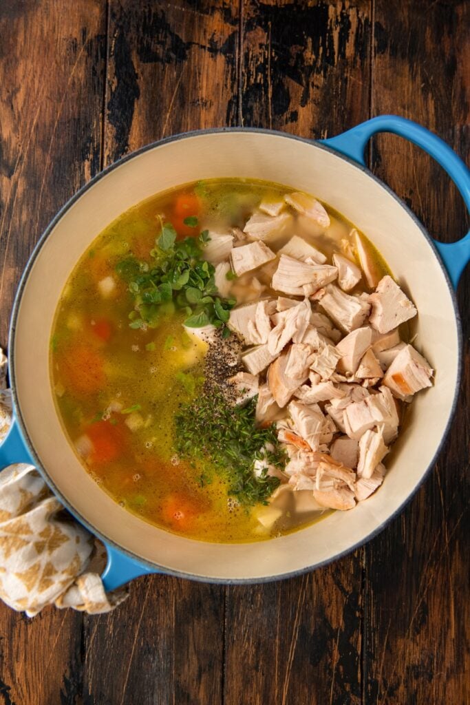 A blue pot filled with homemade chicken soup on a rustic wooden table, containing chopped rotisserie chicken, carrots, celery, herbs, and broth, with a patterned towel next to the handle.