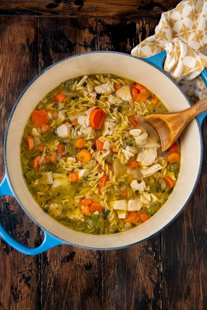 A pot of soup with carrots, celery, chunks of chicken, and uncooked noodles sits on a wooden table. A wooden spoon rests in the soup, and a patterned cloth is nearby. The pot is blue with two handles.