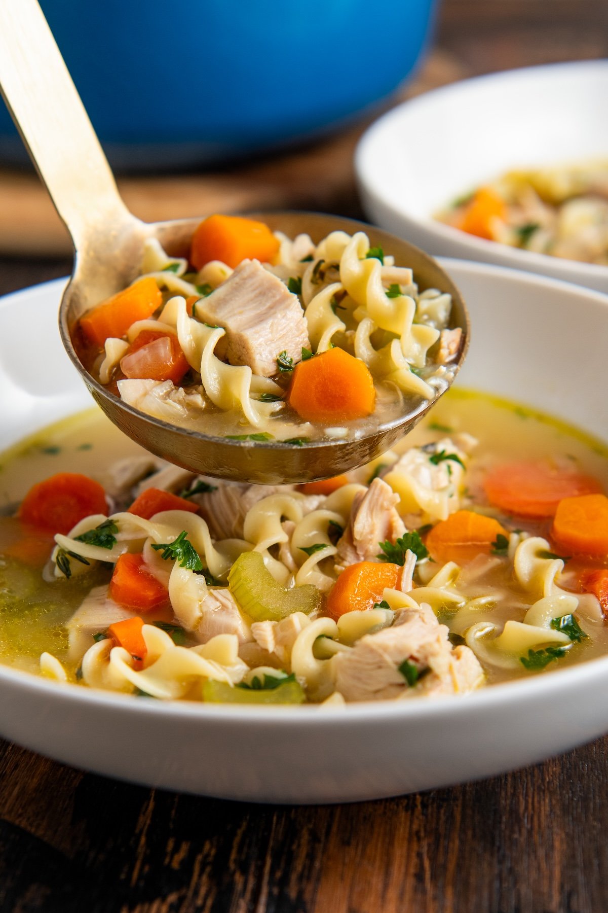 A ladle of rotisserie chicken noodle soup with carrots, celery, egg noodles, and herbs being served into a white bowl, with another bowl and a blue pot in the background.