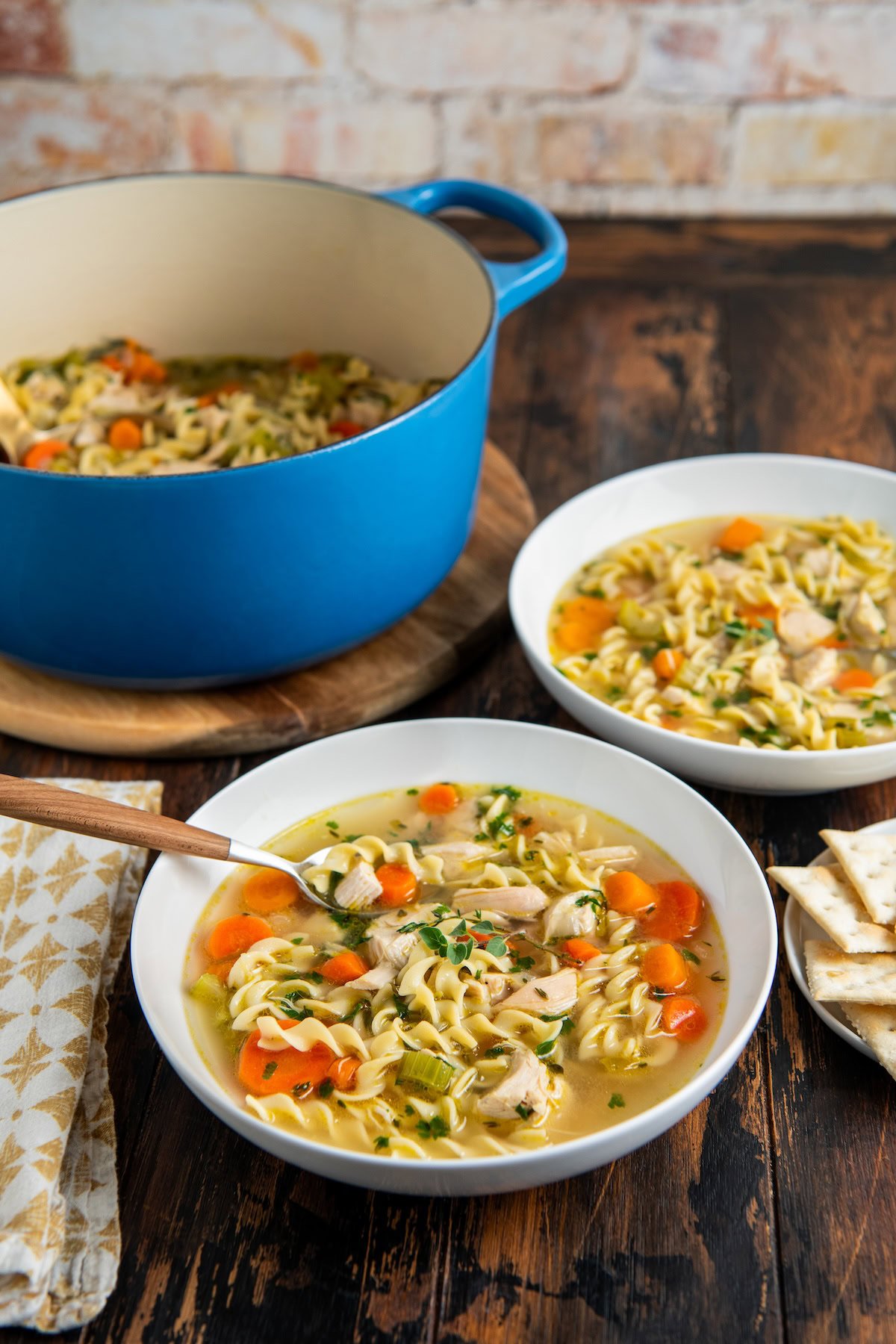 A blue pot and two white bowls filled with rotisserie chicken noodle soup, containing pasta, carrots, celery, and herbs, are arranged on a wooden table with a napkin, spoon, and crackers nearby.