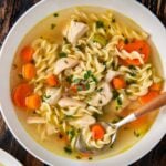 A bowl of chicken noodle soup with rotisserie chicken, egg noodles, sliced carrots, celery, herbs, and chunks of chicken, served with a spoon on a rustic wooden table.