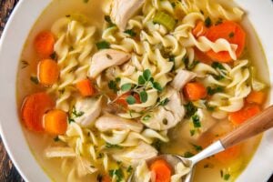 A bowl of chicken noodle soup with rotisserie chicken, egg noodles, sliced carrots, celery, herbs, and chunks of chicken, served with a spoon on a rustic wooden table.