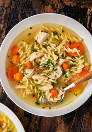A bowl of chicken noodle soup with rotisserie chicken, egg noodles, sliced carrots, celery, herbs, and chunks of chicken, served with a spoon on a rustic wooden table.