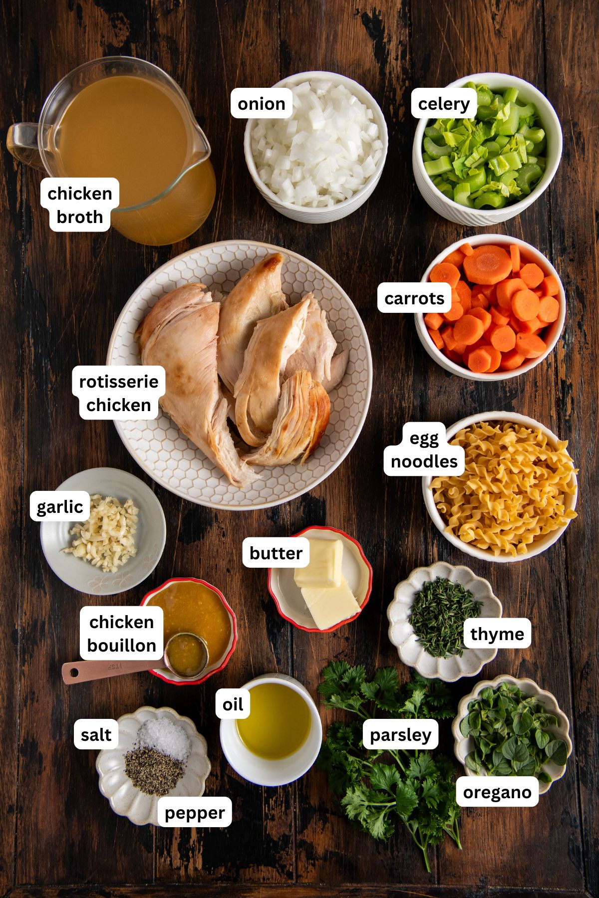 Ingredients for chicken noodle soup with rotisserie chicken arranged on a wooden table, including labeled bowls of chopped onion, celery, carrots, cooked chicken, egg noodles, garlic, parsley, thyme, oregano, chicken broth, bouillon, butter, oil, salt, and pepper.