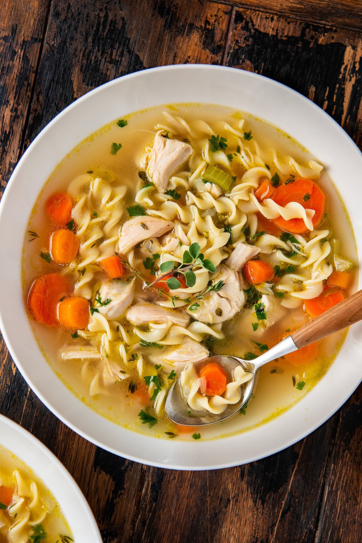 A bowl of chicken noodle soup with rotisserie chicken, egg noodles, sliced carrots, celery, herbs, and chunks of chicken, served with a spoon on a rustic wooden table.