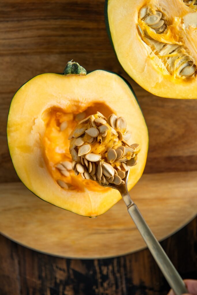 A close-up of an acorn squash cut in half on a wooden surface, with a spoon scooping out seeds and pulp from the center of one half.