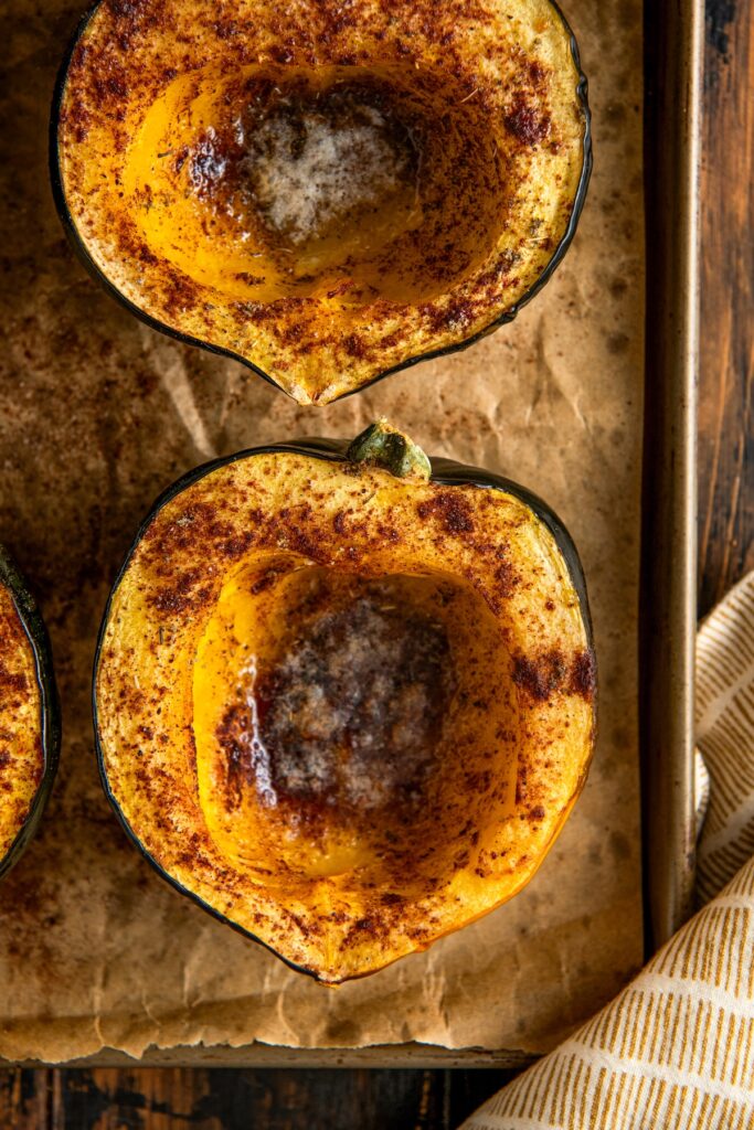 Two baked acorn squash halves on a parchment paper lined baking sheet, sprinkled with cinnamon and a pat of melted butter in the center of each.