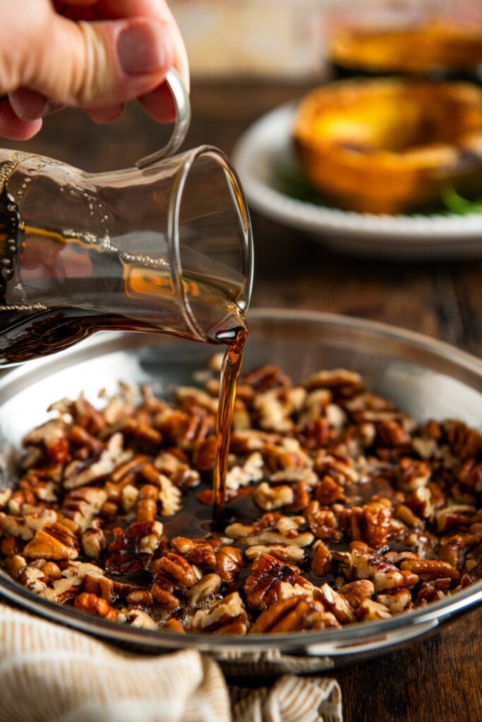 A hand pours a maple syrup from a glass pitcher onto a pan filled with chopped pecans.