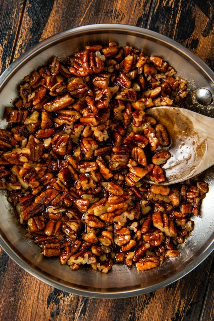 A pan filled with glazed, caramelized maple pecans being stirred with a wooden spoon on a rustic wooden surface.