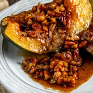 A baked acorn squash half topped with a maple pecans glaze sits on a white plate.