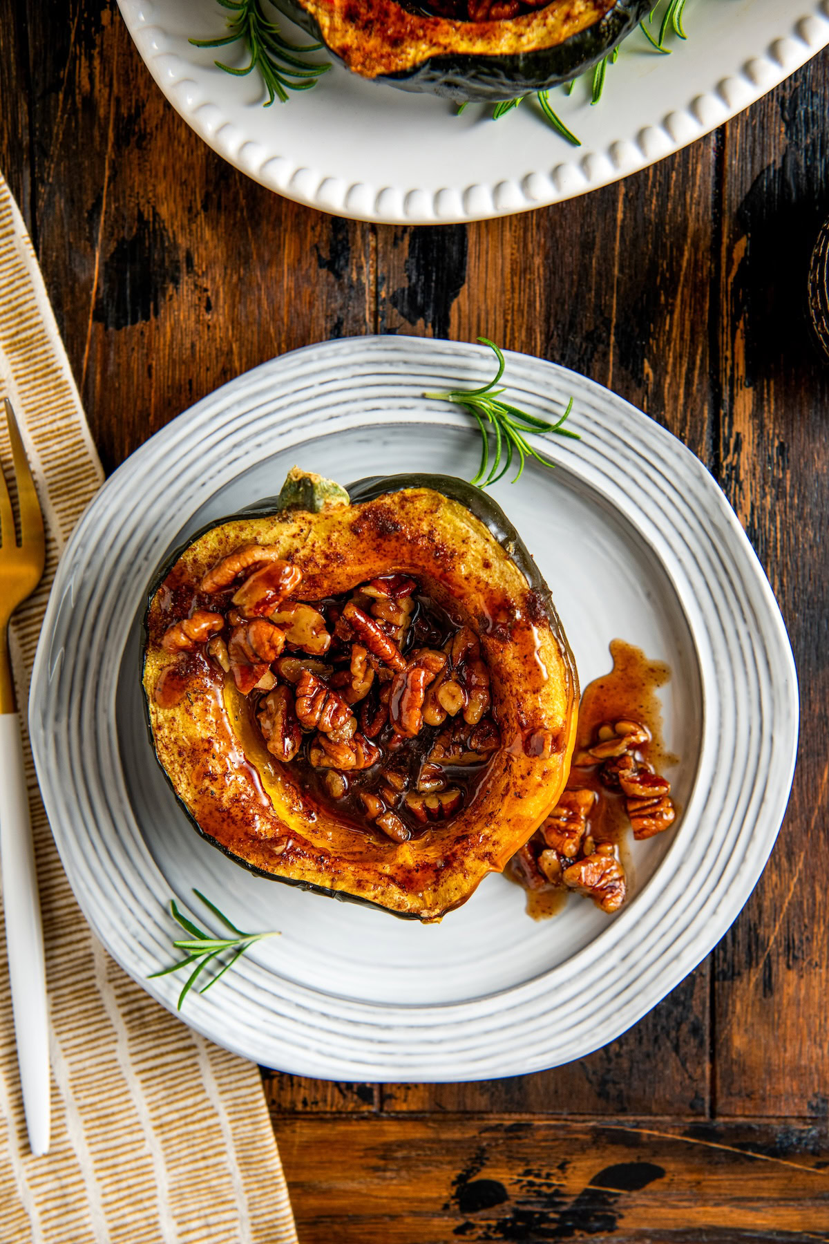 A halved baked acorn squash filled with roasted pecans and maple glaze sits on a white plate, garnished with a sprig of rosemary.