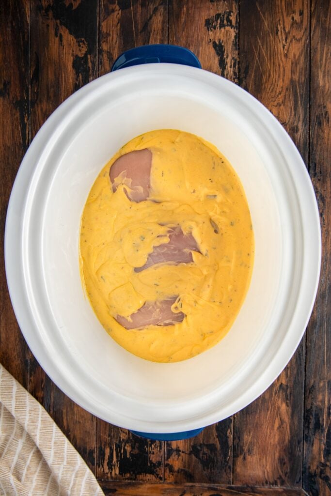 A white oval slow cooker contains raw chicken breasts covered in a thick layer of yellow sauce.
