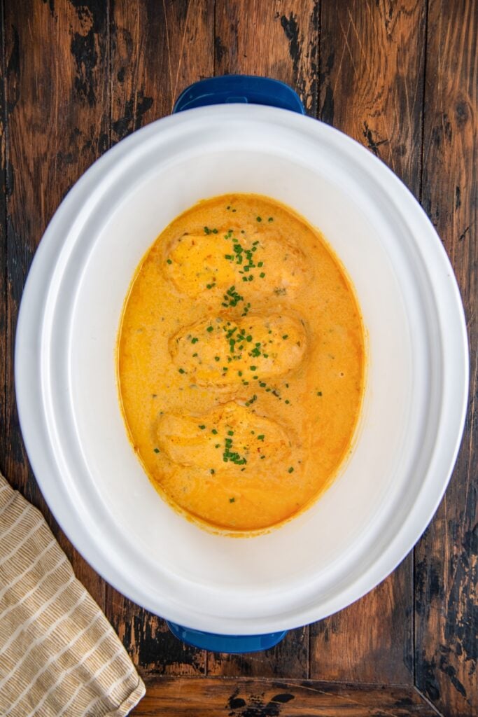 A white slow cooker filled with creamy orange sauce and three pieces of cooked chicken, garnished with chopped chives.
