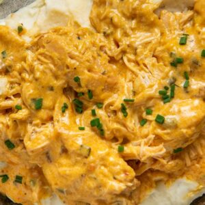 A plate of creamy mashed potatoes topped with shredded French Market chicken in a rich orange sauce, garnished with chopped chives.