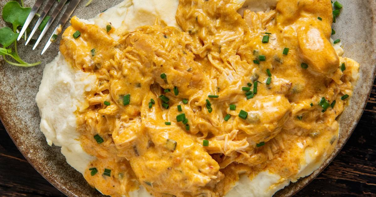A plate of creamy mashed potatoes topped with shredded French Market chicken in a rich orange sauce, garnished with chopped chives.