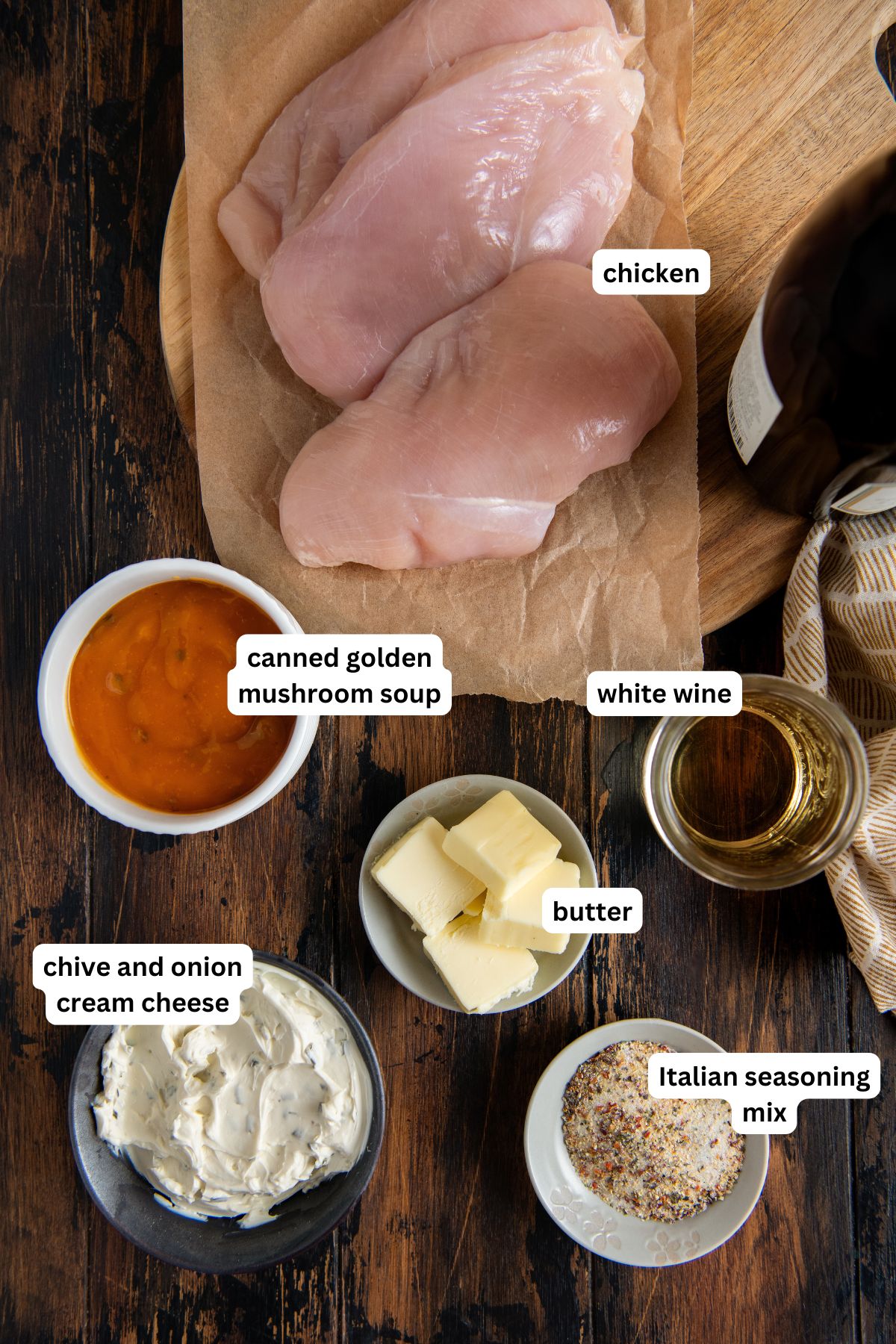 Ingredients for crockpot French Market chicken: raw chicken breasts on parchment, a bowl of canned golden mushroom soup, a small glass of white wine, several cubes of butter, a bowl of chive and onion cream cheese, and Italian seasoning mix on a wooden table.