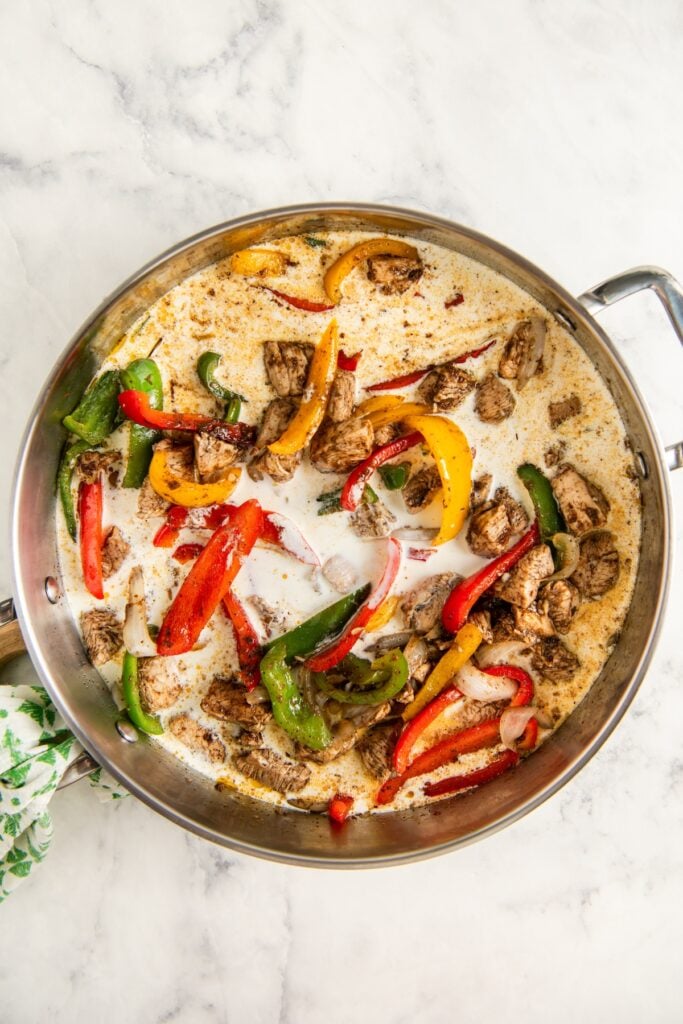 A stainless steel skillet filled with sliced red, yellow, and green bell peppers, onions, and chunks of cooked jerk chicken with heavy cream poured in the middle, sitting on a marble countertop.