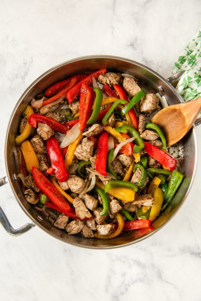 A stainless steel skillet filled with cooked jerk chicken pieces, red, yellow, and green bell peppers, and onions, with a wooden spoon resting on the side.