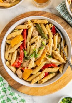 A bowl of rasta pasta with jerk chicken, red and yellow bell peppers, green onions, and creamy sauce, served on a wooden board with a spoon.