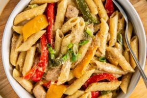 A bowl of rasta pasta with jerk chicken, red and yellow bell peppers, green onions, and creamy sauce, served on a wooden board with a spoon.