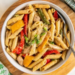 A bowl of rasta pasta with jerk chicken, red and yellow bell peppers, green onions, and creamy sauce, served on a wooden board with a spoon.