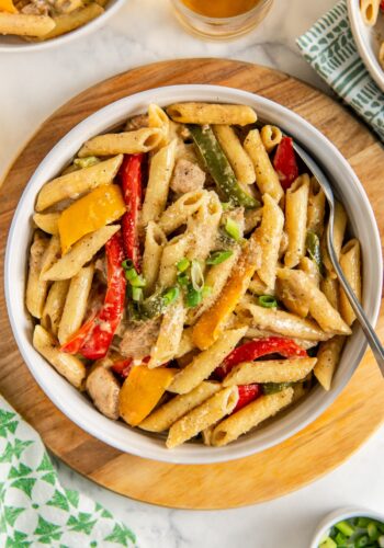 A bowl of rasta pasta with jerk chicken, red and yellow bell peppers, green onions, and creamy sauce, served on a wooden board with a spoon.