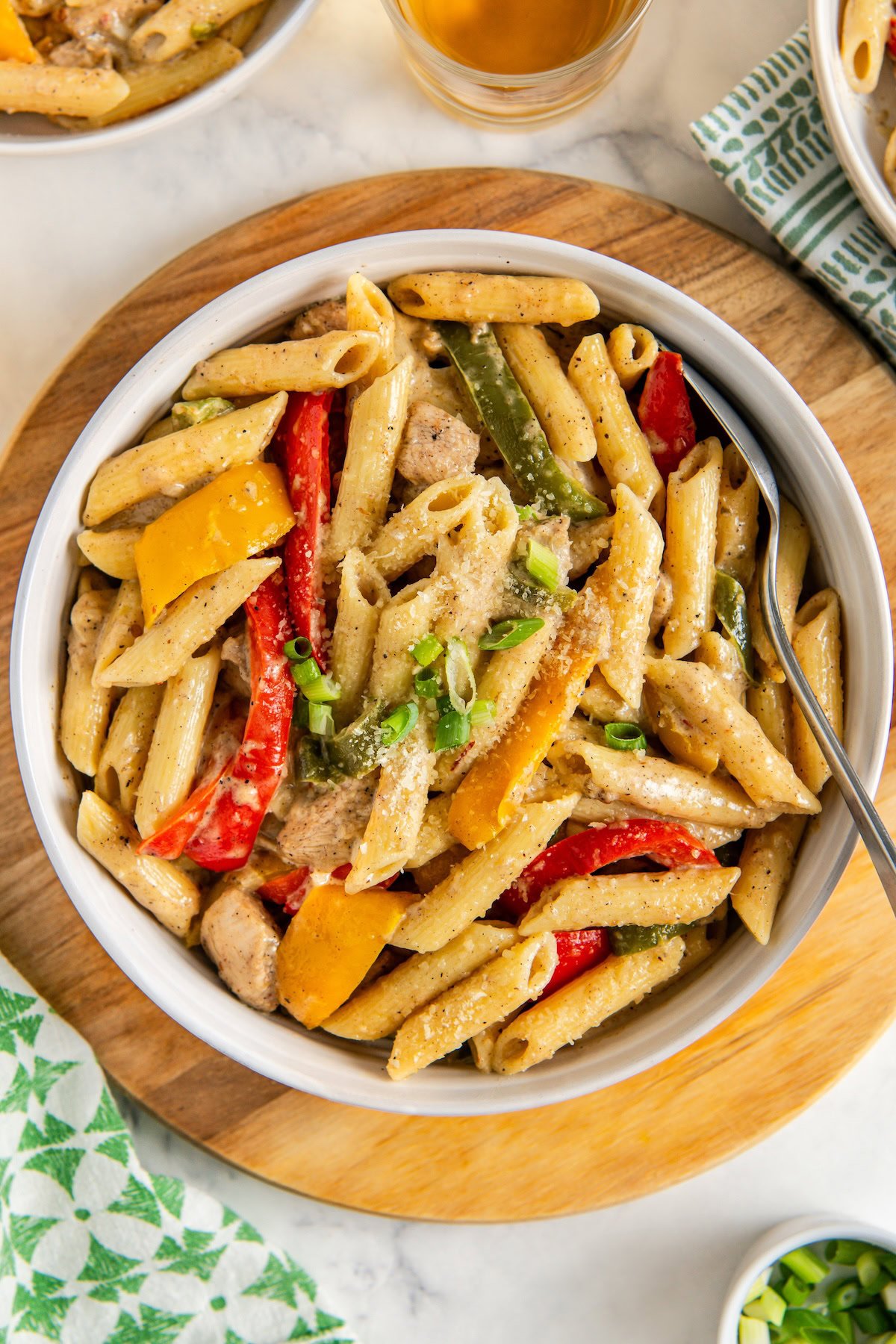 A bowl of rasta pasta with jerk chicken, red and yellow bell peppers, green onions, and creamy sauce, served on a wooden board with a spoon.