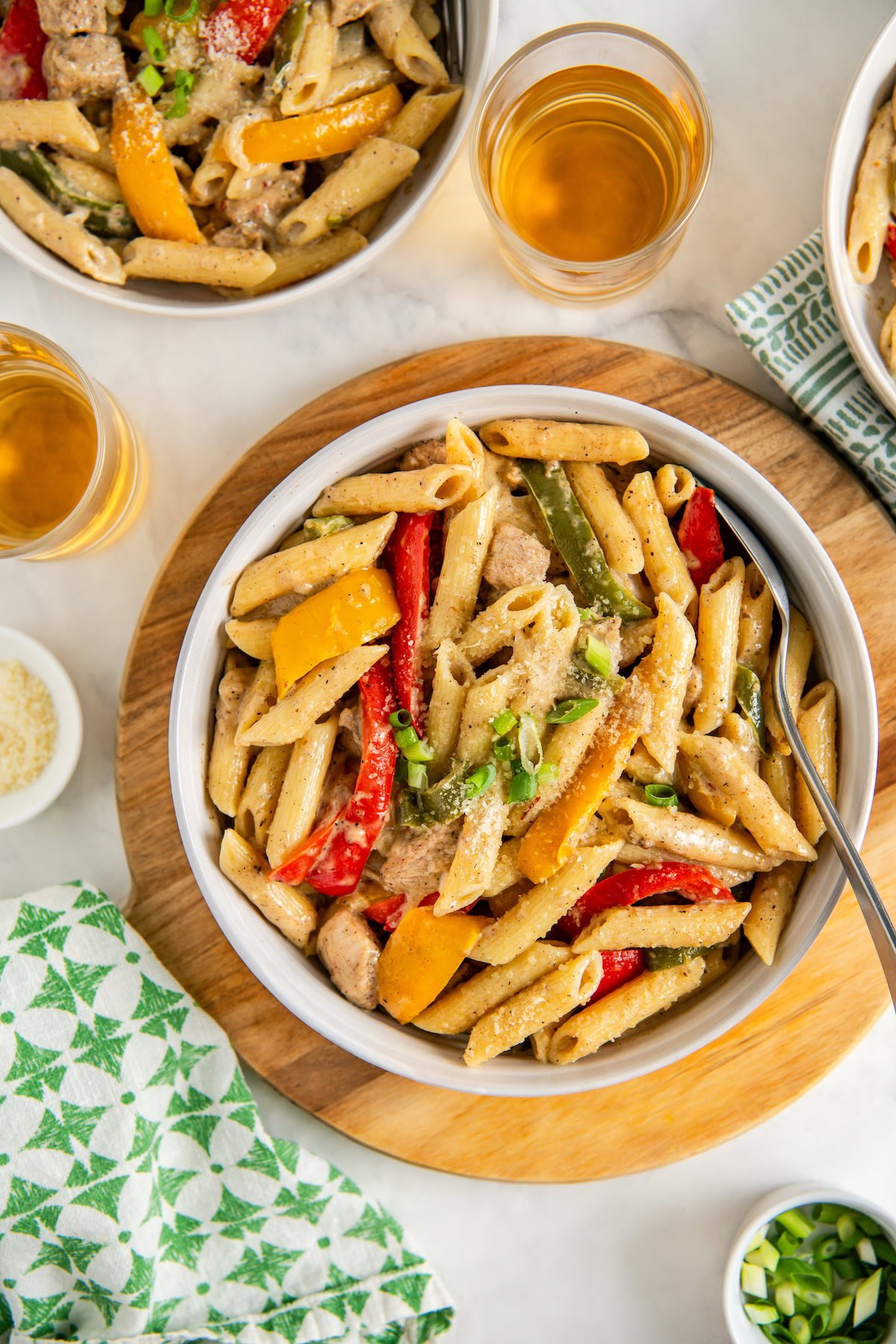A bowl of creamy rasta pasta with sliced red, yellow, and green bell peppers and pieces of jerk chicken, topped with grated cheese, served on a wooden board with two drink glasses nearby.