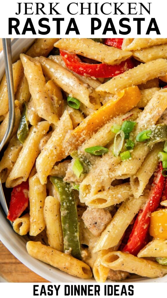 Close-up of a bowl of creamy rasta pasta with jerk chicken, red and yellow bell peppers, green onions, and sprinkled parmesan cheese. A fork is placed in the dish.