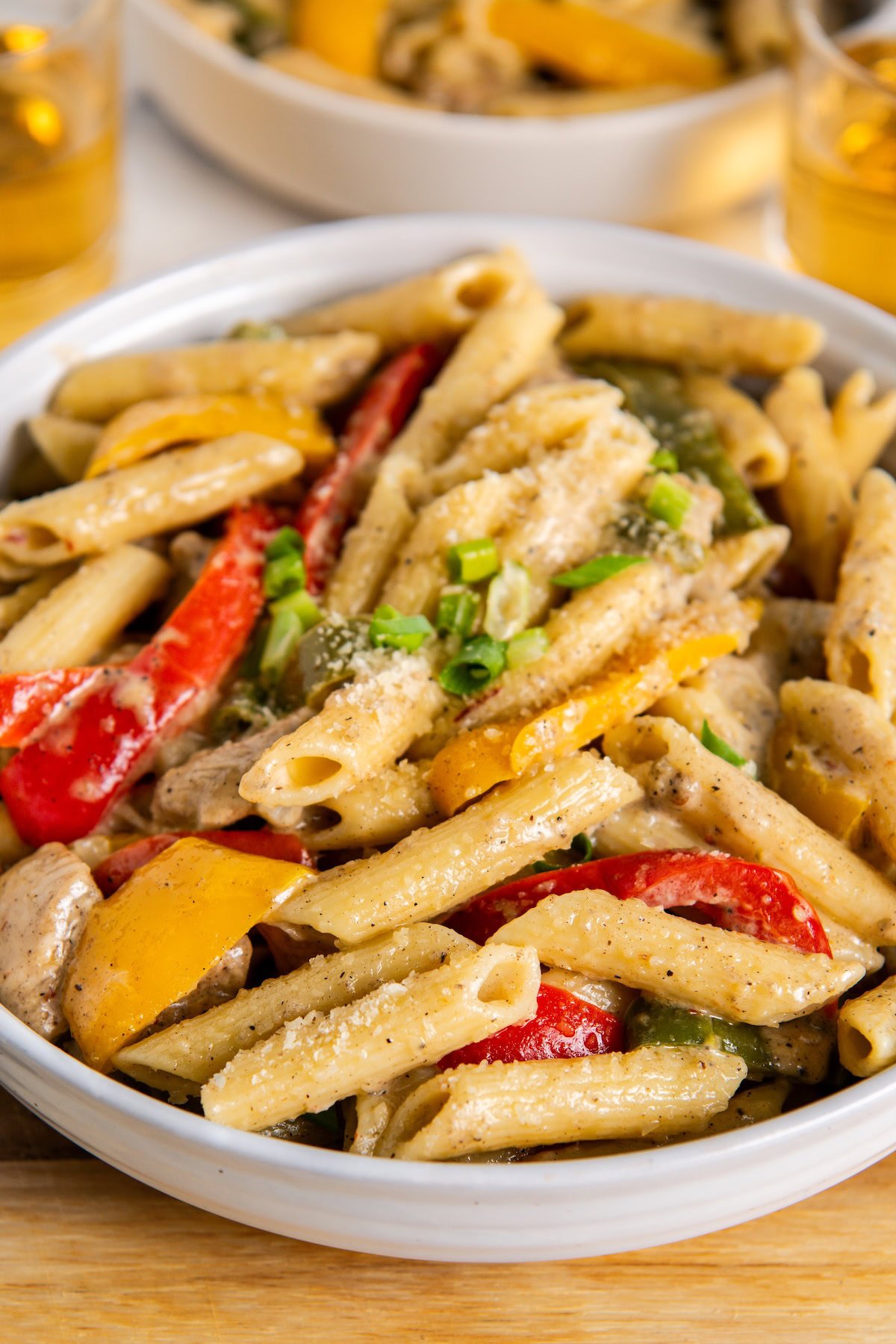 A bowl of jerk chicken rasta pasta with creamy sauce, mixed with red and yellow bell peppers, green onions, and sprinkled with grated cheese.