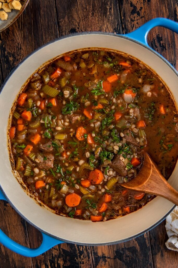 Made easy in one-pot, Beef Barley Soup is loaded with tender beef, chewy pearl barley, and lots of vegetables in a rich and savory broth. A pot filled with beef and barley vegetable soup sits on a wooden table. The soup contains chunks of beef, carrots, celery, barley, and fresh herbs, with a wooden spoon resting in the pot.