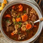 A bowl of beef barley soup with chunks of beef, sliced carrots, barley, and herbs in a rich broth, served on a rustic plate with oyster crackers on the side. A spoon rests in the bowl.