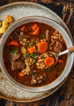 A bowl of beef barley soup with chunks of beef, sliced carrots, barley, and herbs in a rich broth, served on a rustic plate with oyster crackers on the side. A spoon rests in the bowl.