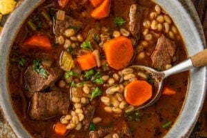 A bowl of beef barley soup with chunks of beef, sliced carrots, barley, and herbs in a rich broth, served on a rustic plate with oyster crackers on the side. A spoon rests in the bowl.