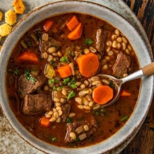 A bowl of beef barley soup with chunks of beef, sliced carrots, barley, and herbs in a rich broth, served on a rustic plate with oyster crackers on the side. A spoon rests in the bowl.