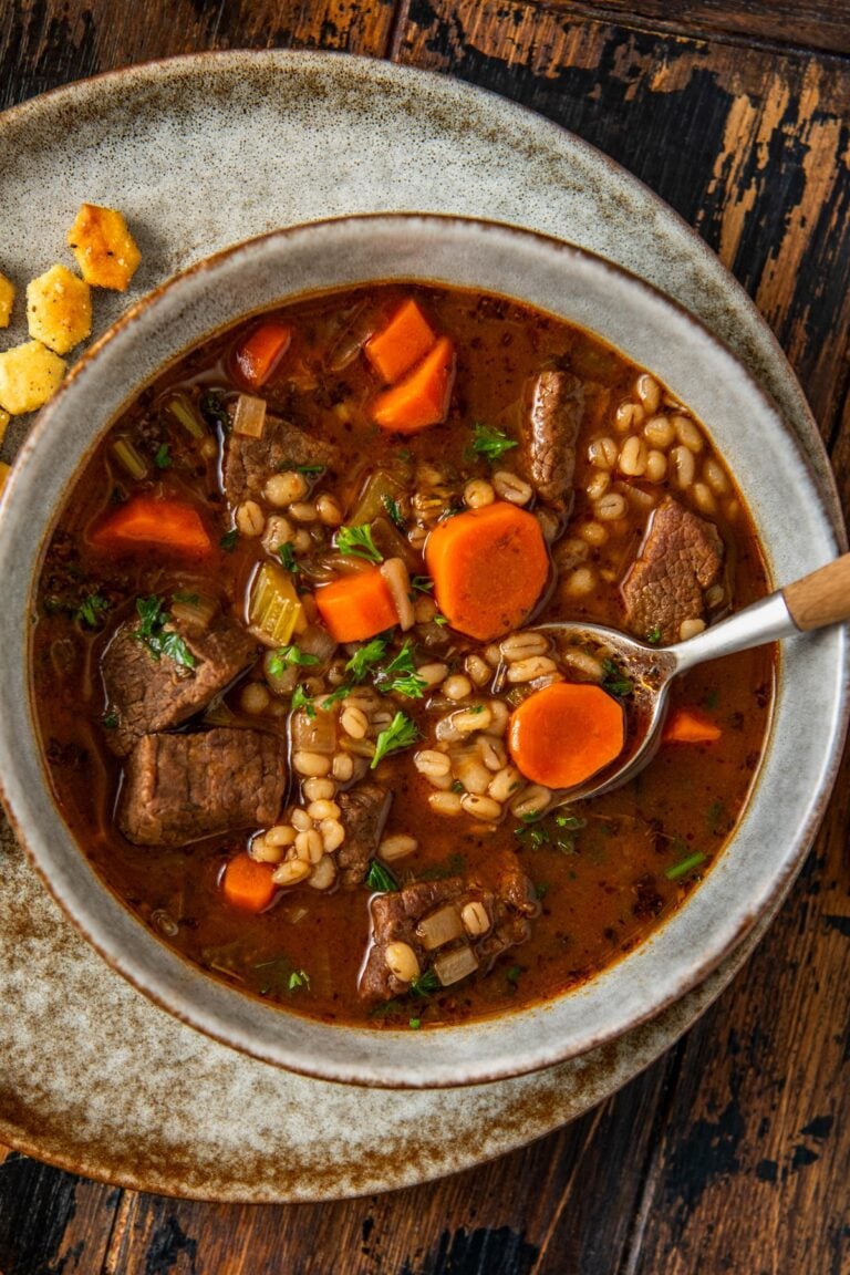 A bowl of beef barley soup with chunks of beef, sliced carrots, barley, and herbs in a rich broth, served on a rustic plate with oyster crackers on the side. A spoon rests in the bowl.