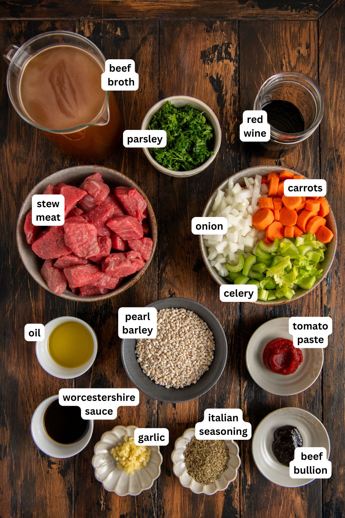 Made easy in one-pot, Beef Barley Soup is loaded with tender beef, chewy pearl barley, and lots of vegetables in a rich and savory broth. View of labeled ingredients for beef barley soup recipe on a wooden table for beef barley soup, including beef broth, stew meat, barley, carrot, celery, onion, parsley, garlic, seasonings, tomato paste, oil, Worcestershire sauce, red wine, and beef bouillon.